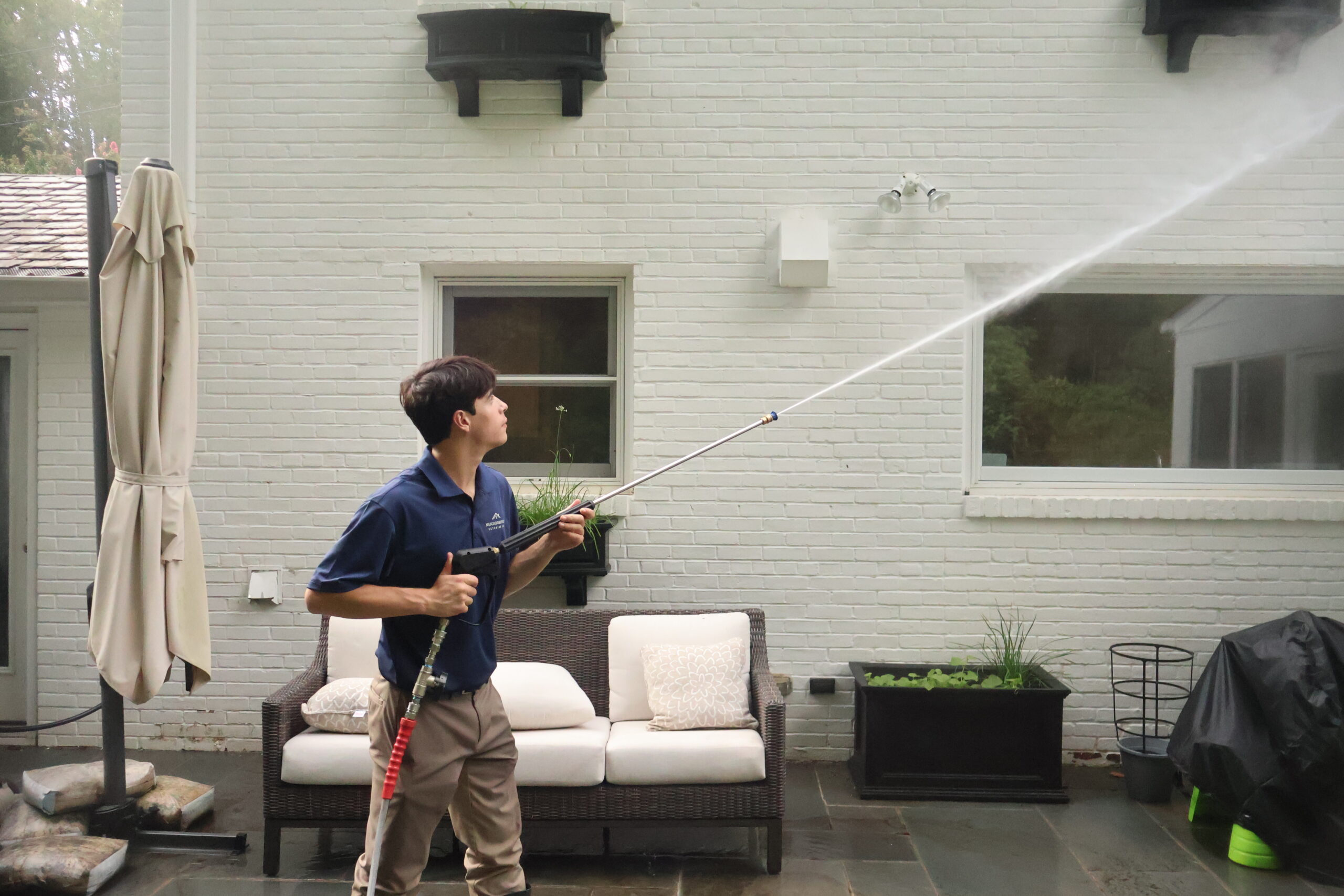 A Neighborhood Exterior technician soft washing a painted brick wall with a long extension wand.